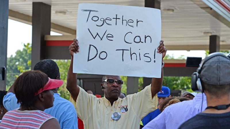 Demonstrator standing with group outdoors holding a handwritten sign saying 