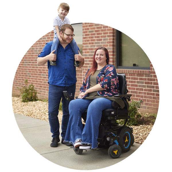 A family consisting of a man, woman, and child. Child is on the standing man's shoulders smiling and woman is sitting in motorized wheelchair.