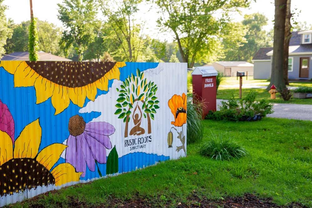 Rustic roots mural painted on a fence outside depicting bright flowers and the logo of a person meditating under a tree arch