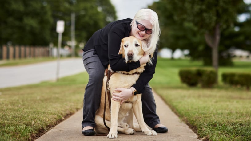 Woman with white hair and dark glasses with vision impairment hugs guide dog on sidewalk