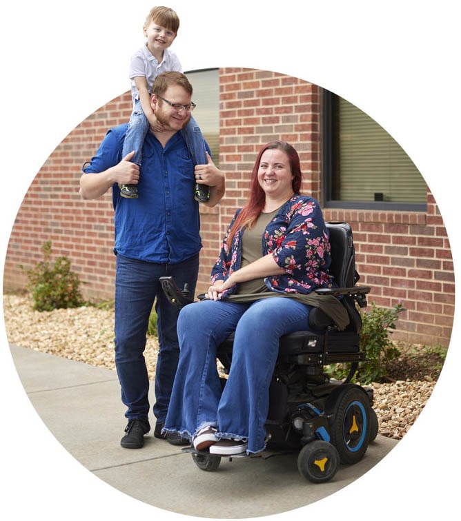 Image of woman in a motorized wheelchair next to standing man with a small child on his shoulders, outdoor on sidewalk in front of brick building.