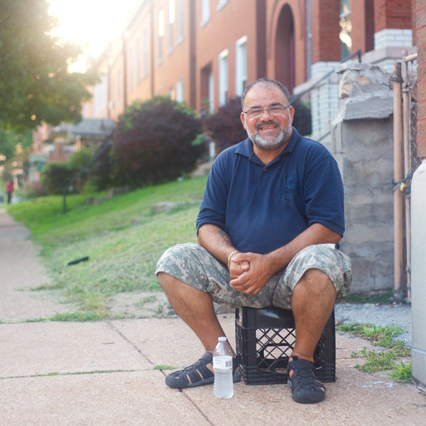 Photo of a Man hanging out on city sidewalk sitting on a milk crate, smiling at the the camera