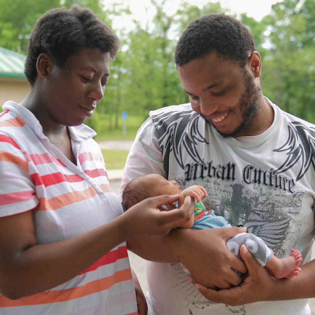 young black parents smile while looking at infant held in father's arms. They are outdoors with trees visible in the background
